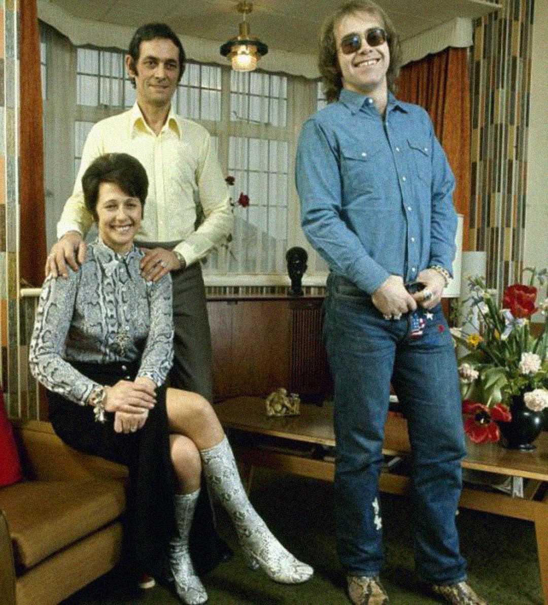 Three adults pose in a living room. A woman sits on a couch wearing a patterned dress and boots, with a man in a suit standing beside her. Another man in sunglasses and denim smiles, standing to the right. Flowers and decor are visible in the background.