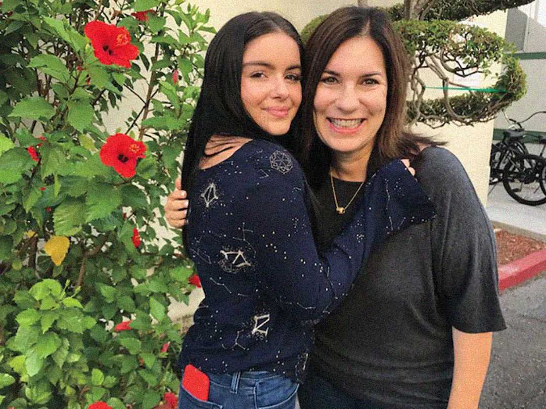 Two women are standing close together, smiling at the camera. One has long dark hair and wears a navy patterned sweater; the other has shoulder-length dark hair and wears a dark top. Red flowers and greenery are behind them.