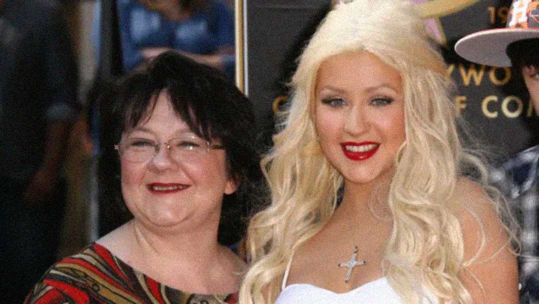 Two women pose together, smiling at the camera. One has long platinum blonde hair and wears a white dress with a cross necklace; the other has short dark hair and glasses, wearing a patterned top.