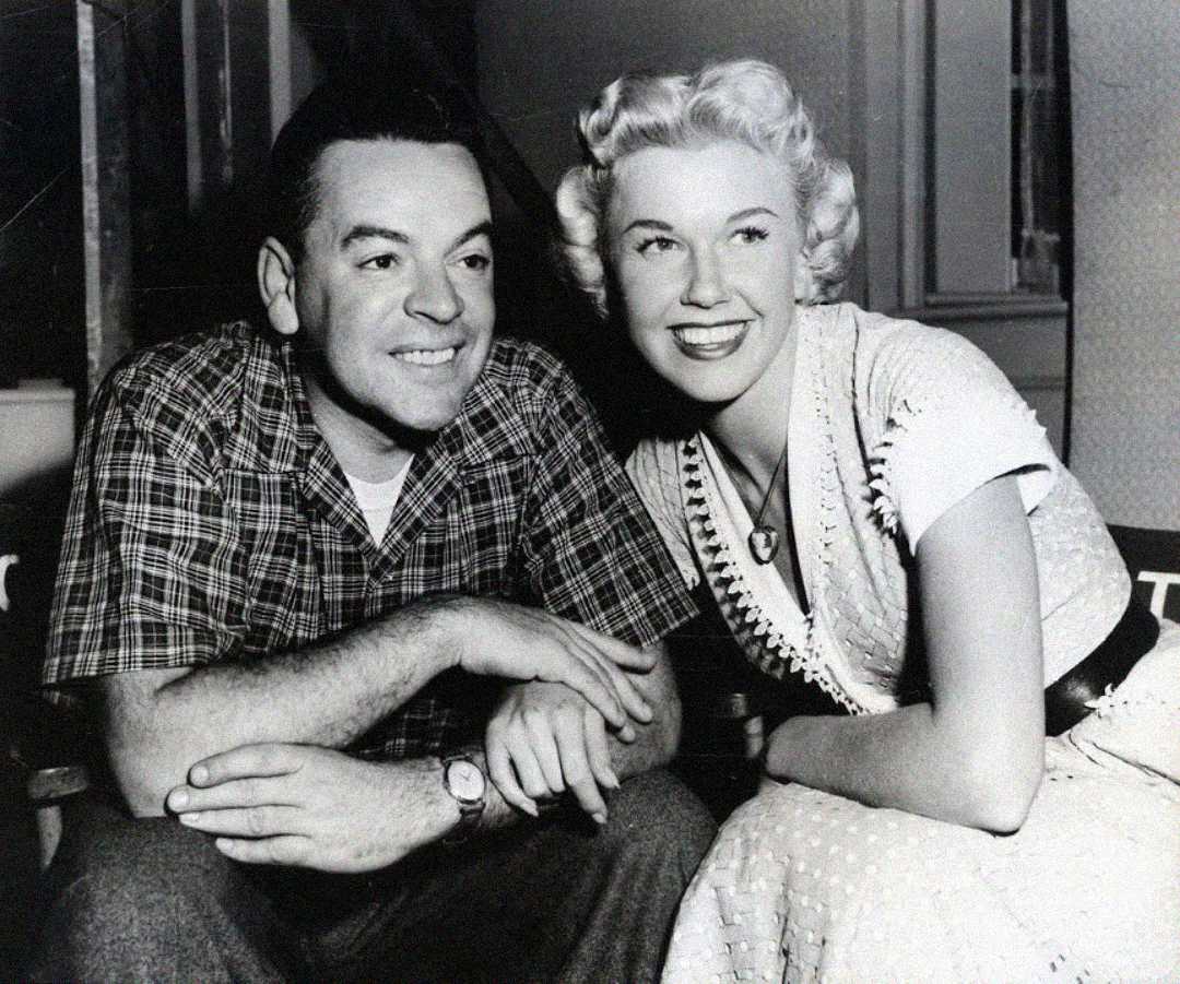 A man in a plaid shirt and a woman in a light-colored dress sit closely together, smiling and looking slightly to the right. The photo appears to be black and white and from the mid-20th century.