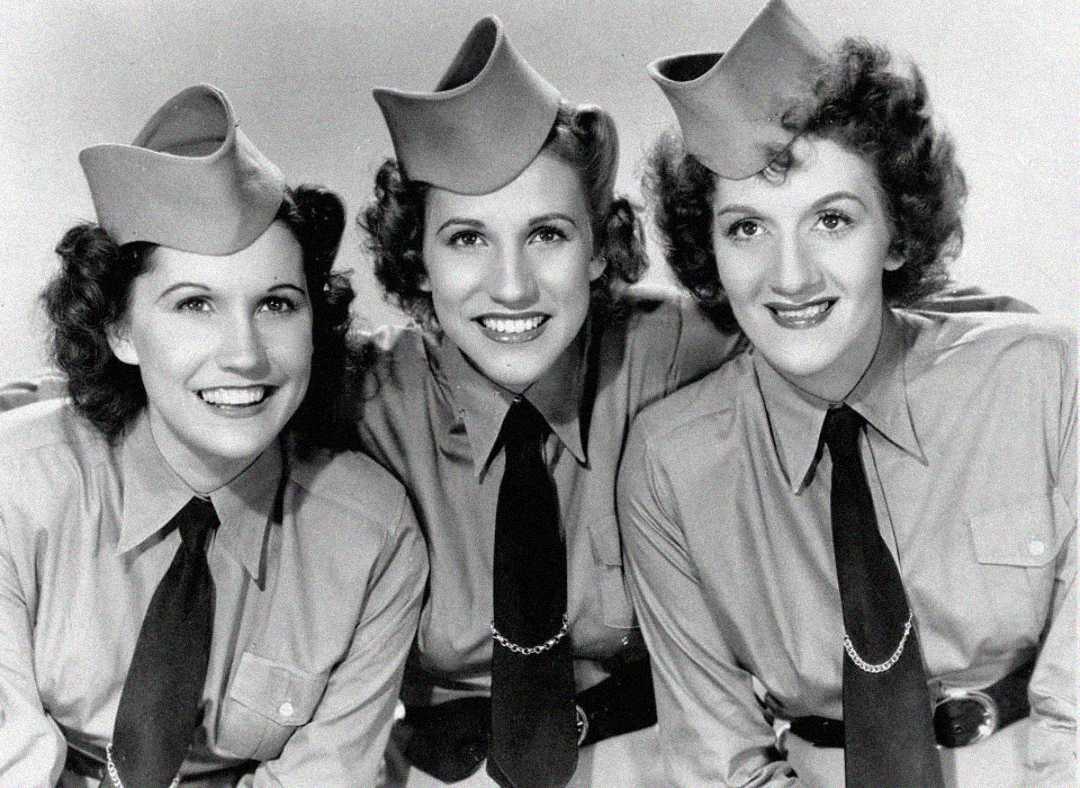 Three women in matching military-style uniforms and hats smile for a black-and-white portrait. Their uniforms feature button-up shirts, ties, and chain details, and their hair is styled in loose curls typical of the 1940s.