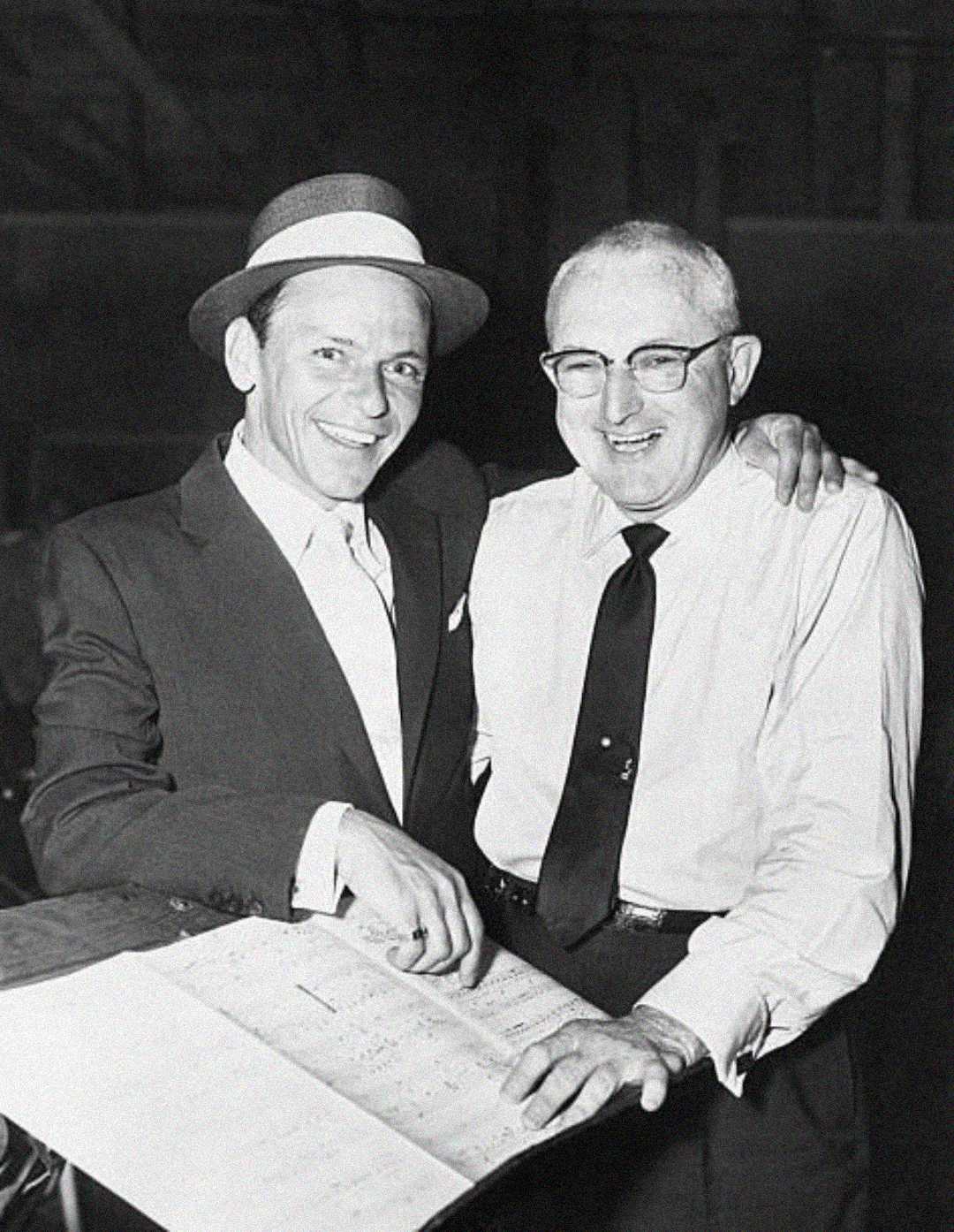 Two men smile while looking at sheet music. The man on the left wears a suit, tie, and hat; the man on the right wears glasses, a white shirt, and a dark tie. They have their arms around each other's shoulders.