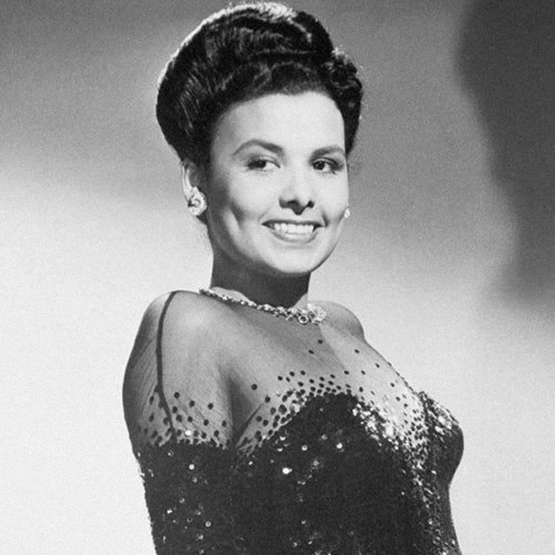 A woman in a glamorous, sequined dress smiles confidently. She has styled, dark hair and wears earrings and a necklace. The photo is black and white with a soft background.