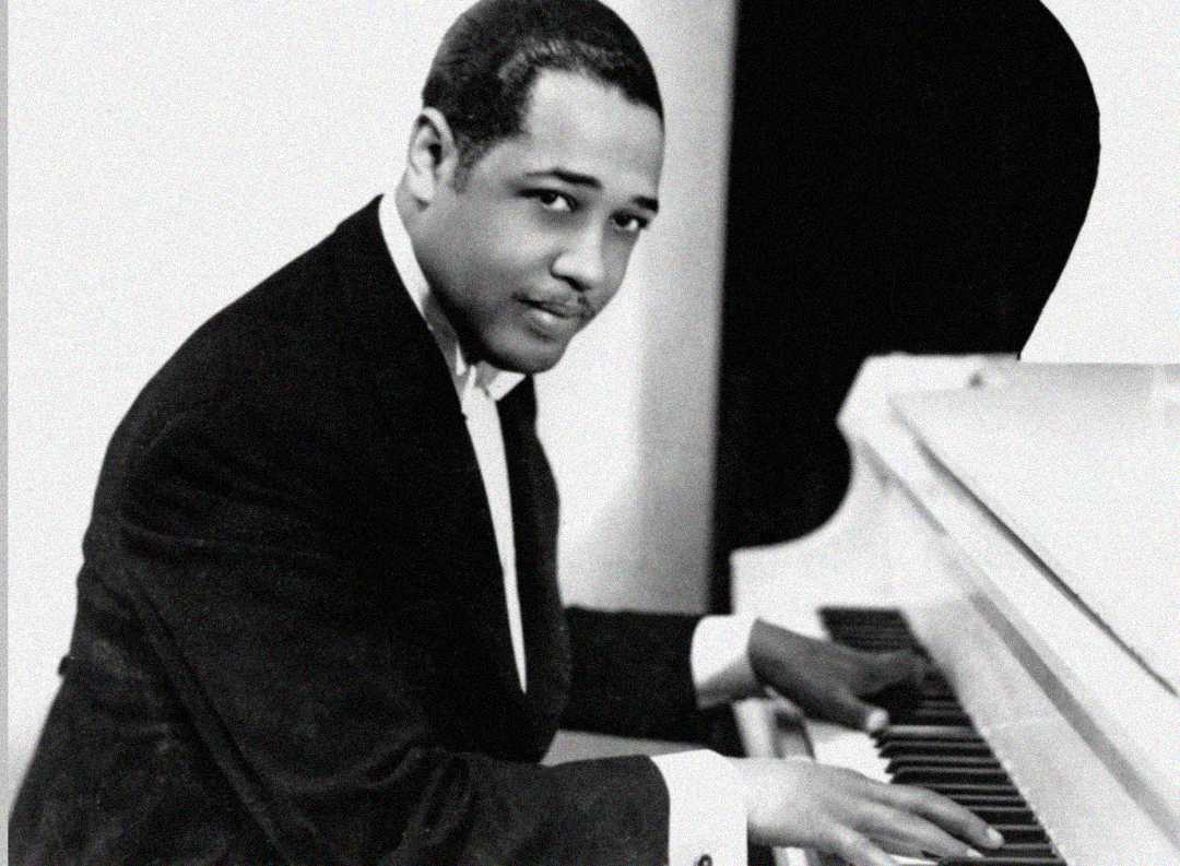 A man in a suit sits at a grand piano, looking toward the camera while playing the keys. The photo is in black and white, giving it a classic, vintage feel.