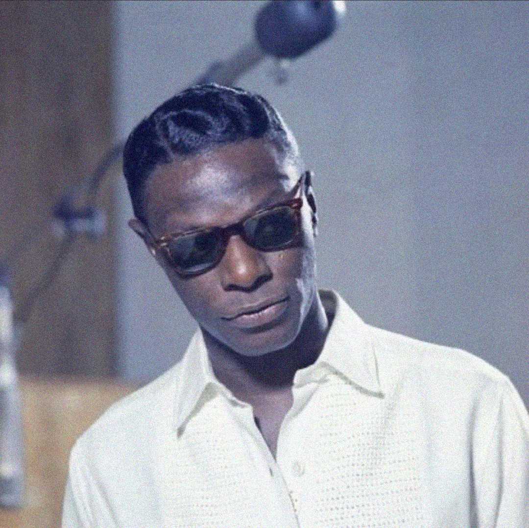 A man wearing dark sunglasses and a light-colored shirt stands indoors, with a large microphone visible in the blurred background.