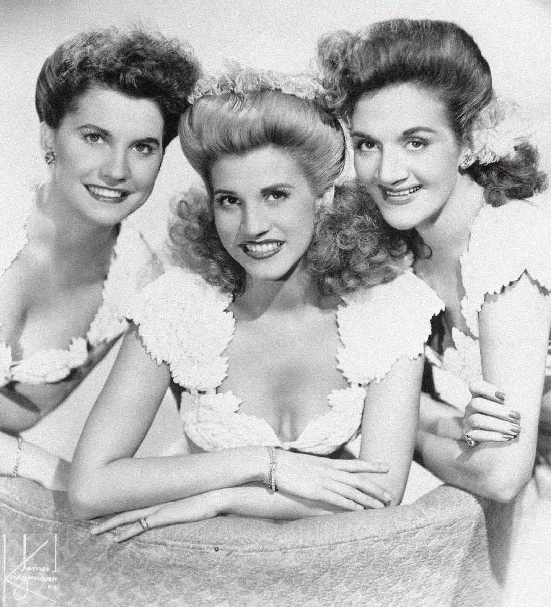 Three women with curled hair and matching white, floral-accented dresses pose closely together, smiling at the camera in a black-and-white vintage studio portrait.