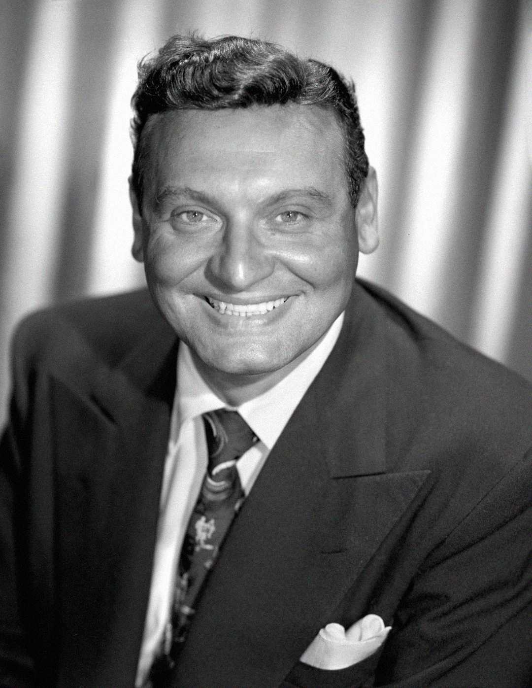 Black and white portrait of a smiling man in a suit and tie, with neatly styled dark hair, posing in front of a softly blurred curtain background.