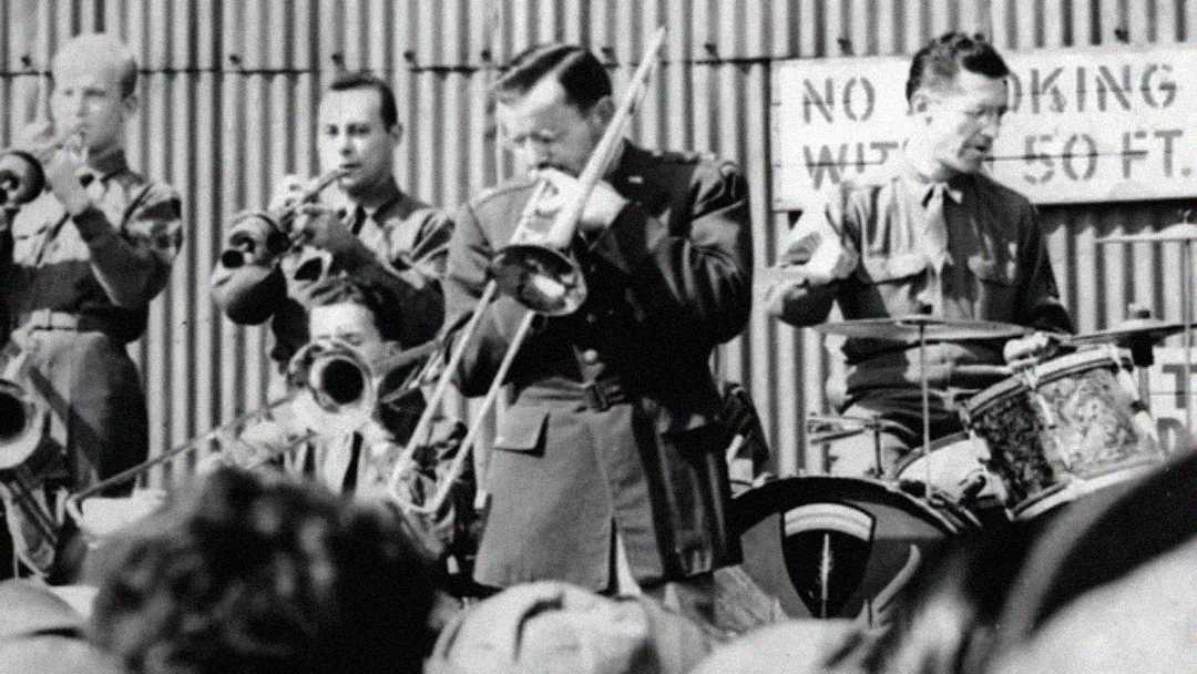 A black-and-white photo of a military band performing outdoors, featuring a trombonist in front, a trumpet player, a drummer, and other musicians. A sign in the background partially reads "NO SMOKING.