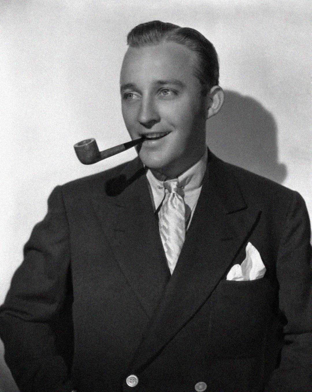 A man in a suit and tie smiles while holding a pipe in his mouth. He has neatly combed hair and a pocket square in his jacket. The photo is black and white with a shadow behind him.