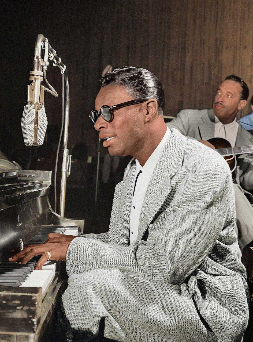 A man in a gray suit and dark sunglasses plays piano and sings into a vintage microphone, with a guitarist sitting behind him, in a dimly lit studio setting.