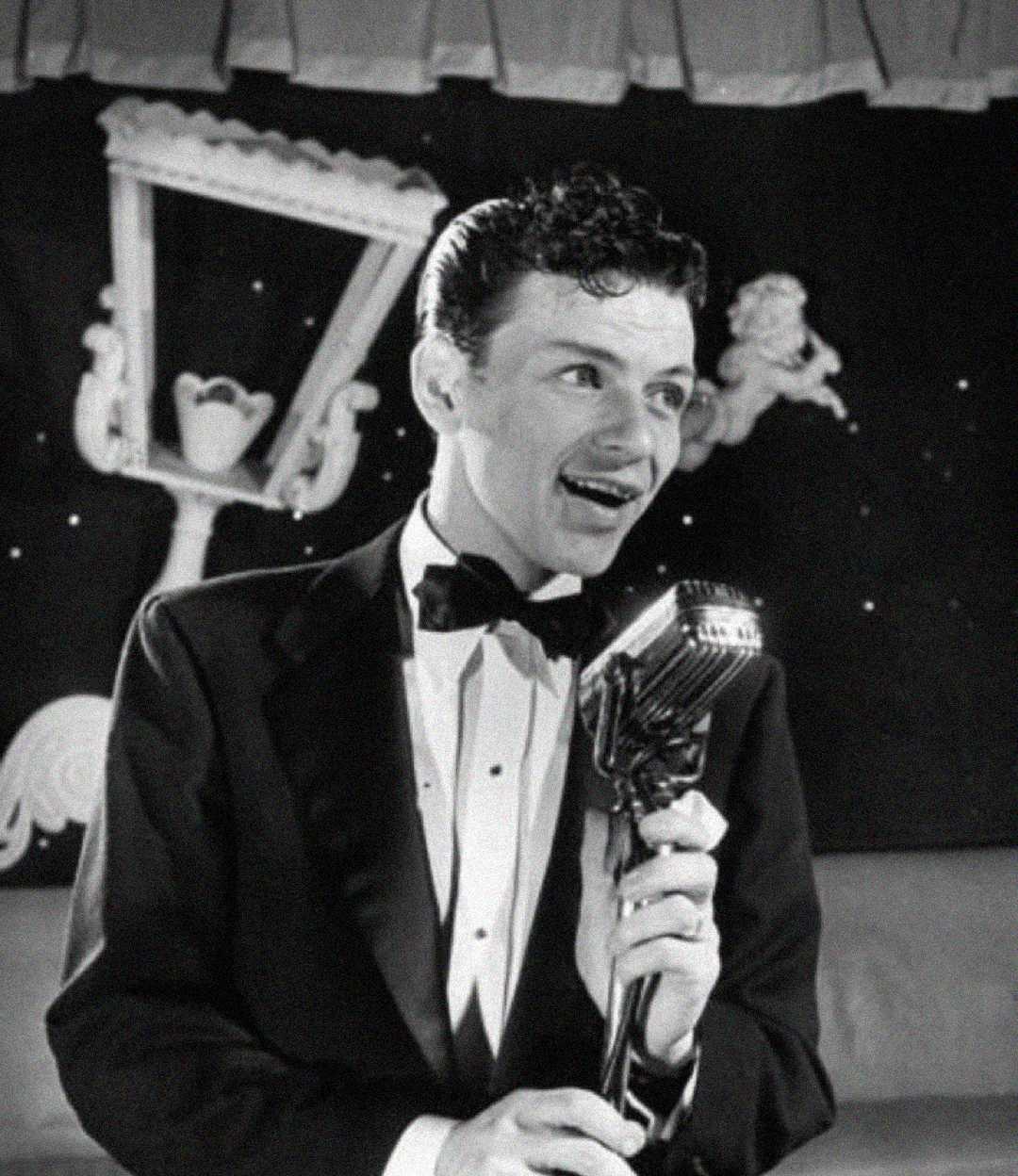 A young man in a tuxedo sings into a vintage microphone with a backdrop featuring artistic designs, including a cherub and an ornate frame.