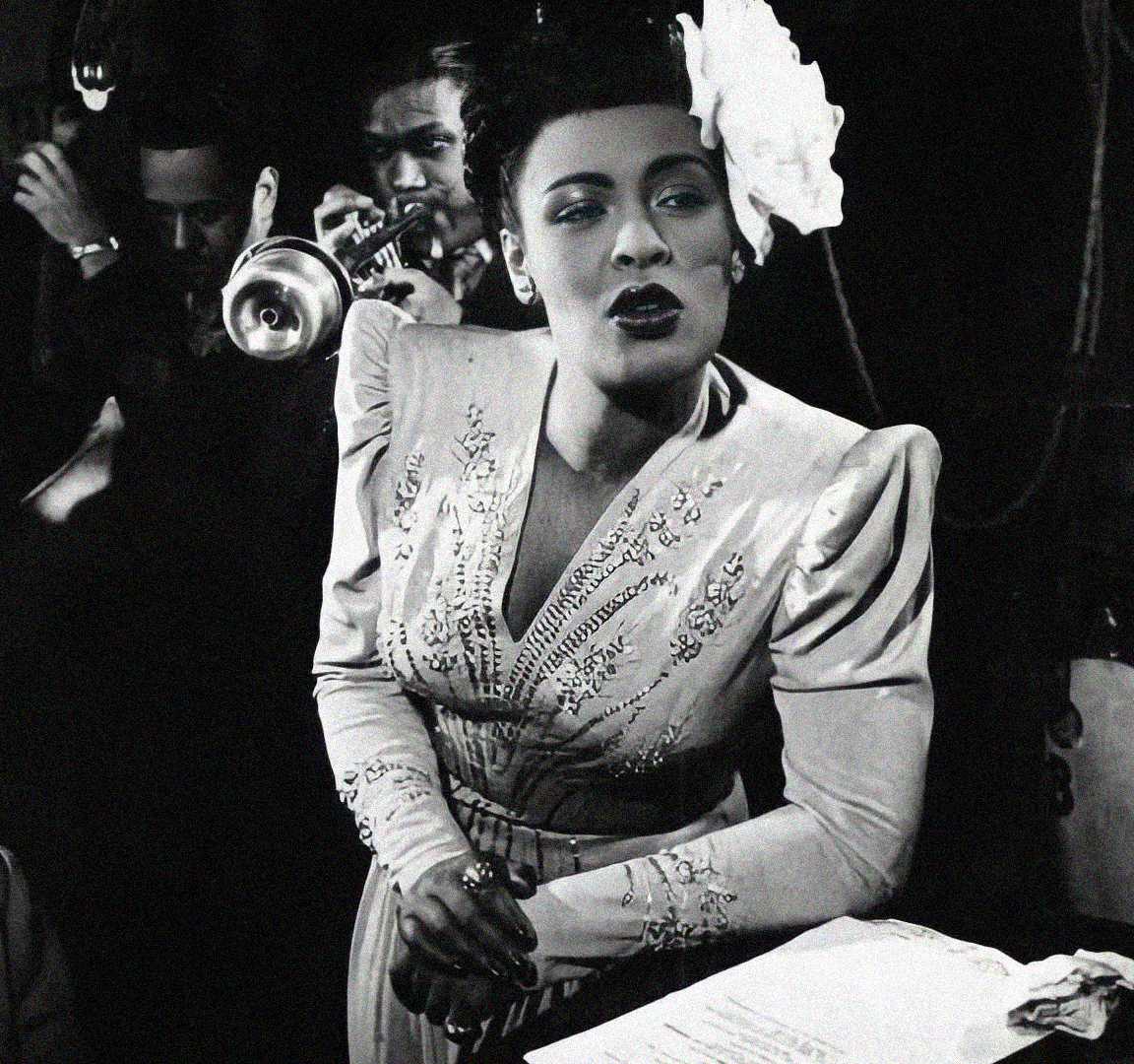 A Black woman in a vintage embellished dress sings passionately into a microphone, with a white flower in her hair. A trumpet player performs in the background in this black-and-white photo.