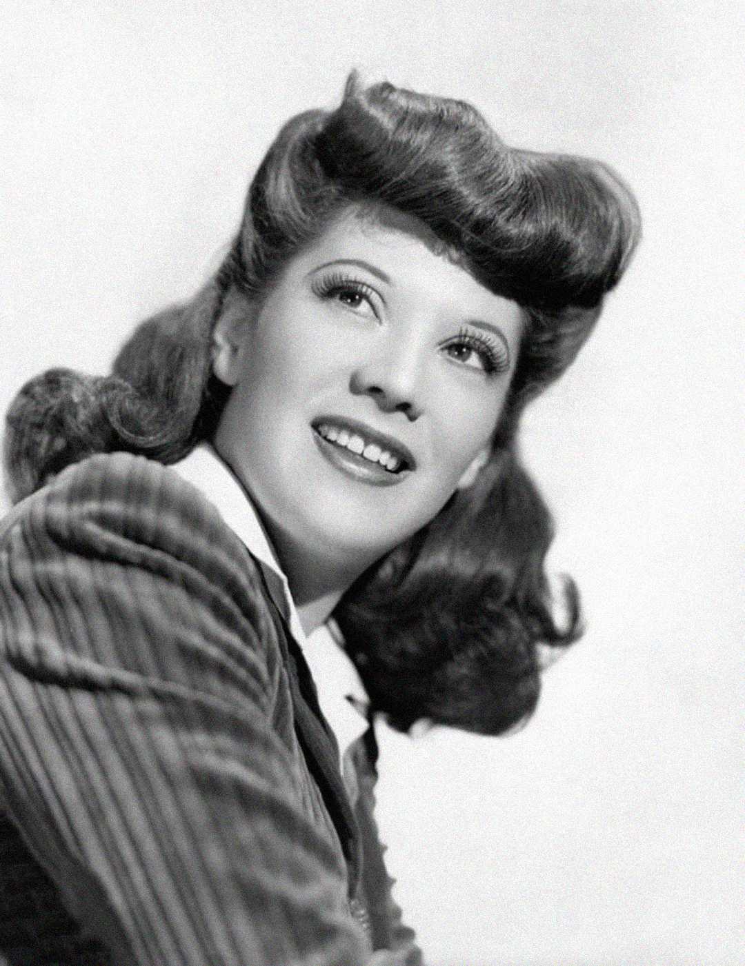 A black and white portrait of a smiling woman with wavy hair styled in vintage waves, wearing a pinstriped jacket and a white collared shirt, looking slightly upward and to the side.