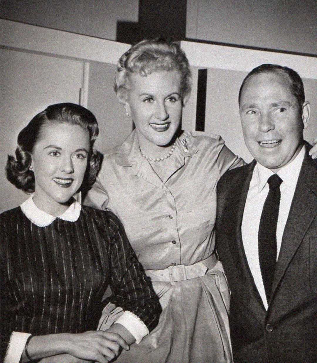 Three people pose together, smiling for the camera. The two women wear vintage dresses and the man wears a suit and tie. The image is in black and white, giving it a classic, mid-20th-century feel.