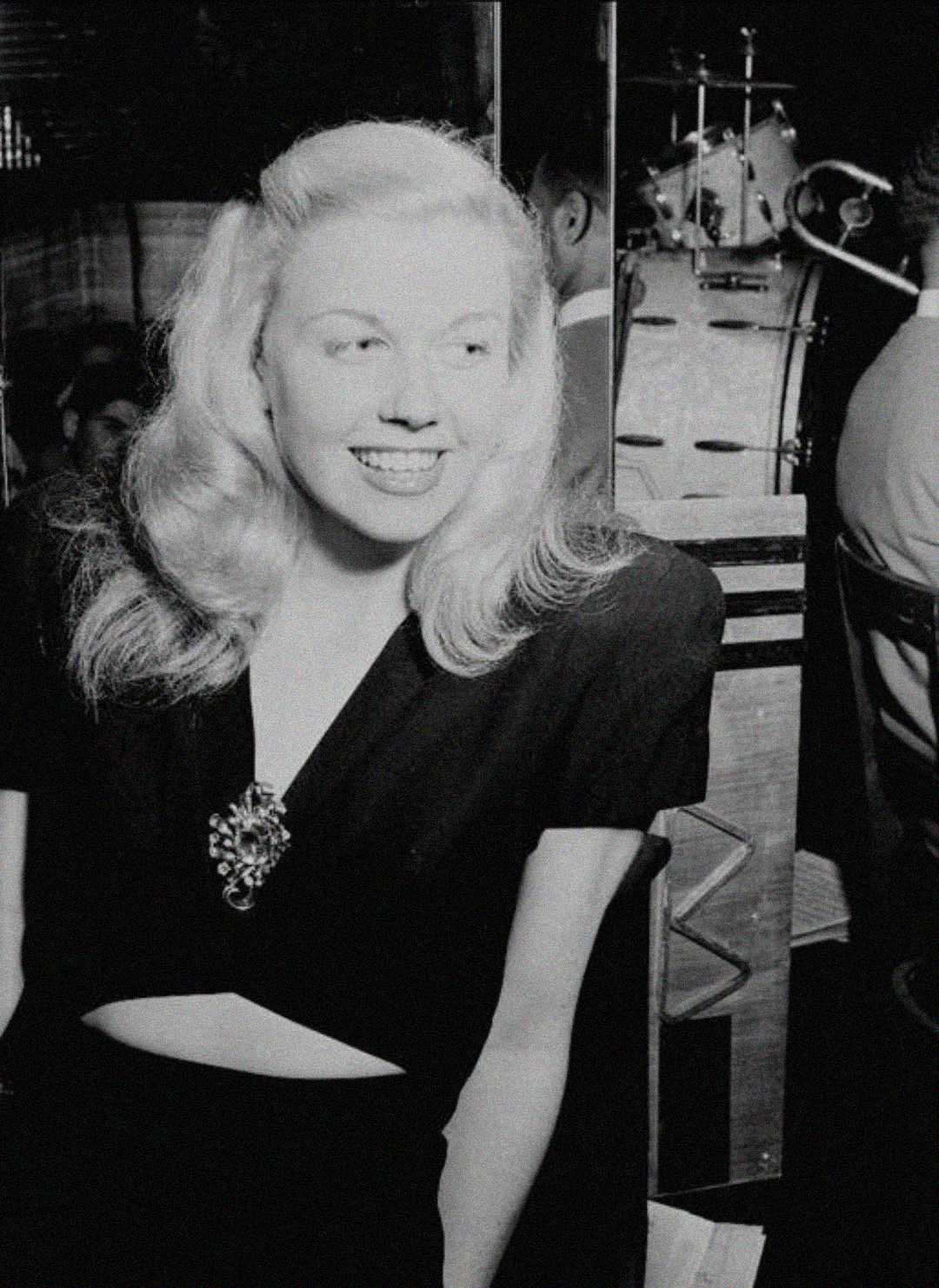 A smiling woman with wavy blonde hair wearing a dark dress stands indoors. In the background, musical instruments and people are visible, suggesting a social or entertainment setting. The image is in black and white.