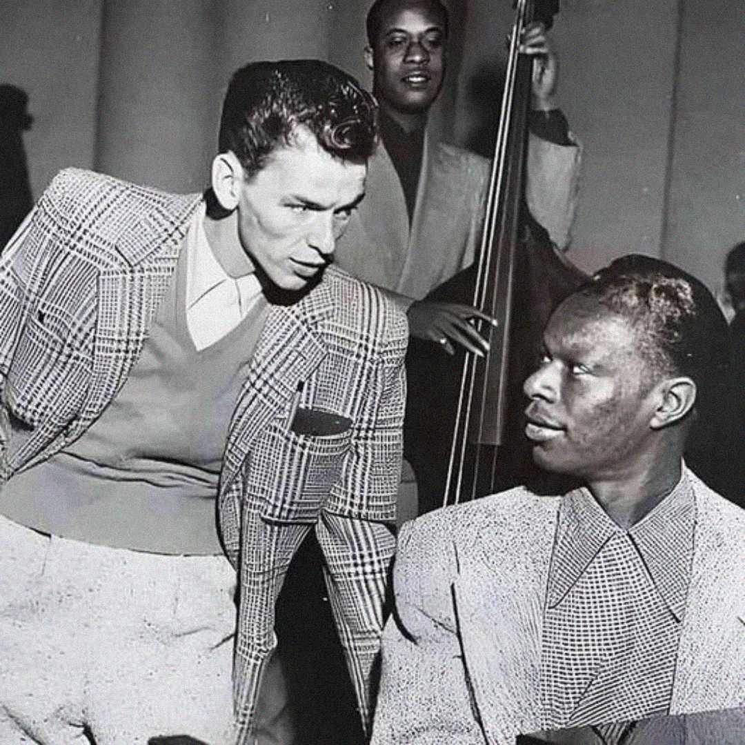 A black-and-white photo shows two men in suits engaged in conversation. One stands and leans forward, while the other sits at a piano. A musician with a double bass is visible in the background.