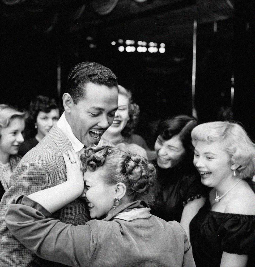 A group of young adults laugh and smile together at a lively social gathering. One woman leans playfully against a man in a suit, while others around them appear to be enjoying the moment. The scene is in black and white.