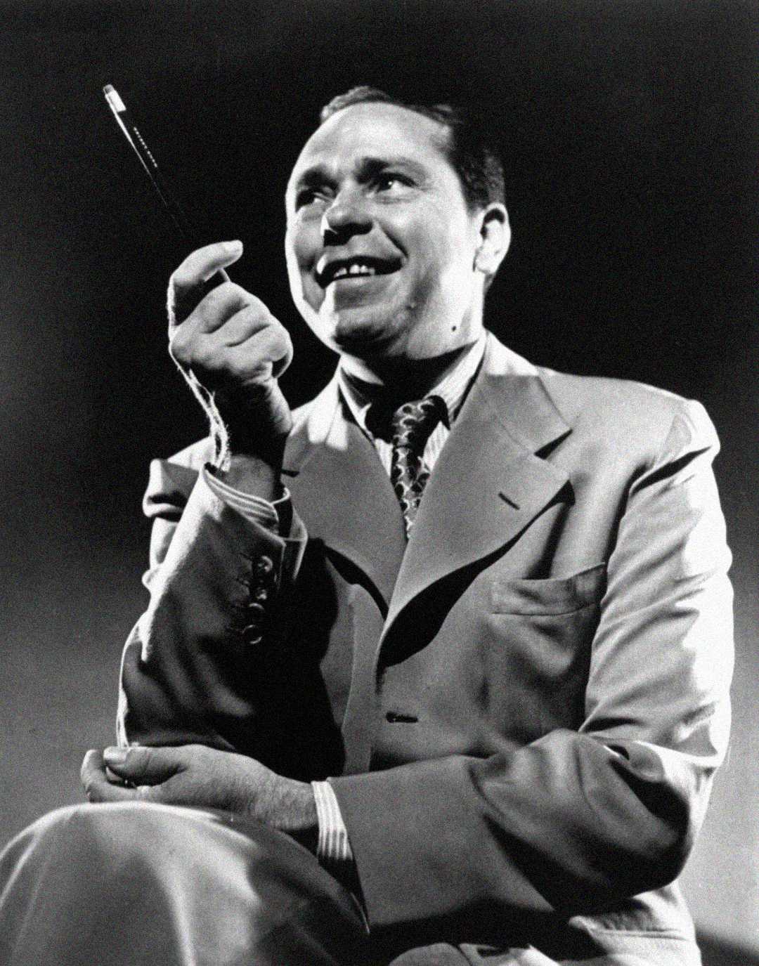 A man in a suit sits with legs crossed, smiling and holding a pen upright in his right hand. The photo is black and white, with dramatic lighting and a dark background.