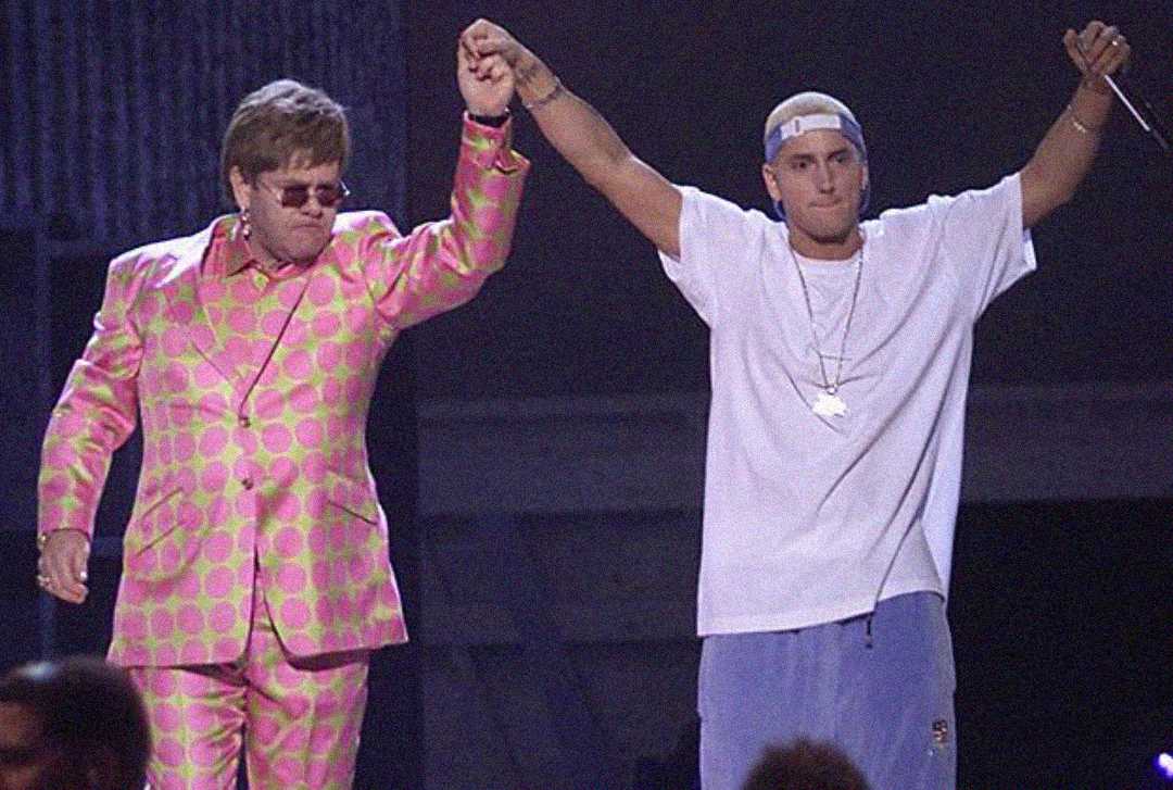 Two men stand on stage holding hands in the air. One wears a vibrant pink and yellow patterned suit with sunglasses; the other wears a white t-shirt, blue pants, and a white cap, holding a microphone.