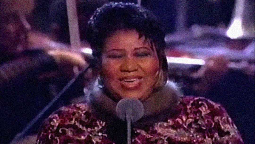 A woman in a patterned dress with a fur collar sings into a microphone, smiling, with musicians and orchestral instruments blurred in the background.