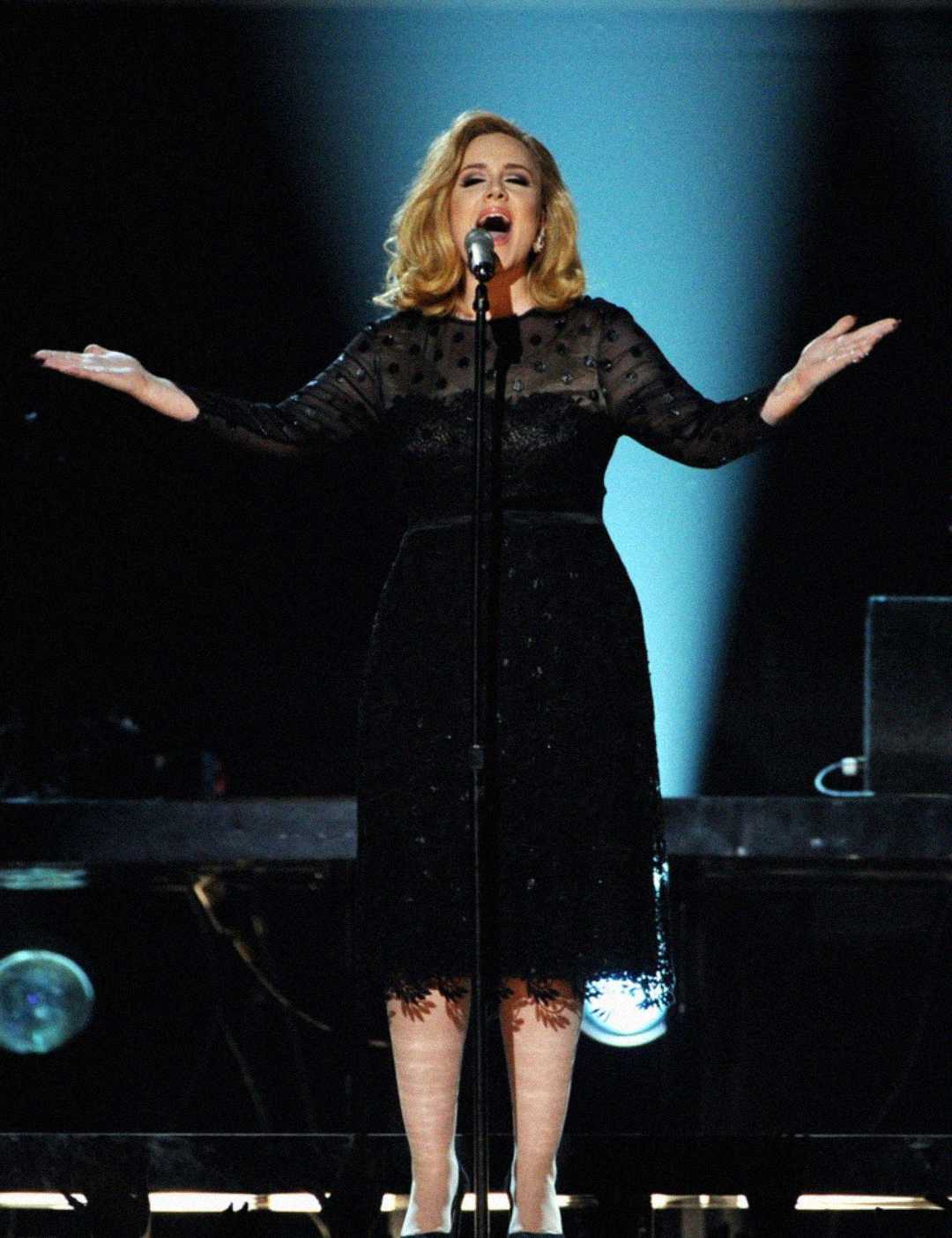A woman in a black dress sings passionately on stage with her arms open wide, standing in front of a microphone and dramatic lighting behind her.