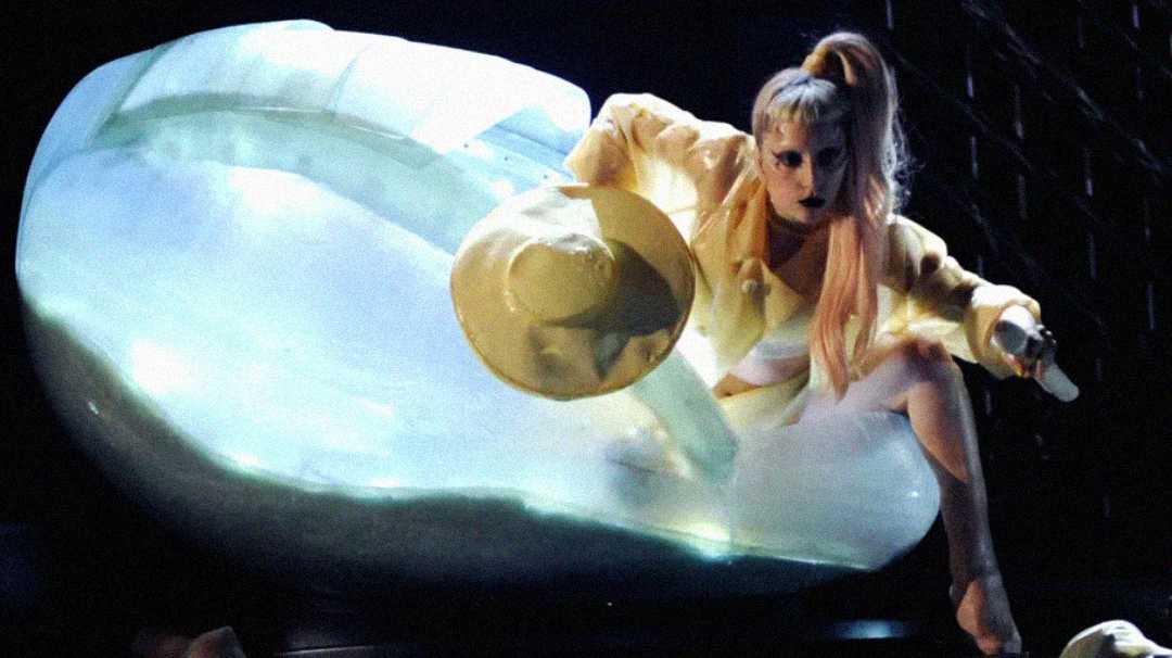 A performer with long blonde hair in a high ponytail, dramatic makeup, and a yellow outfit crouches on a large, glowing clam shell while holding a matching wide-brimmed hat.