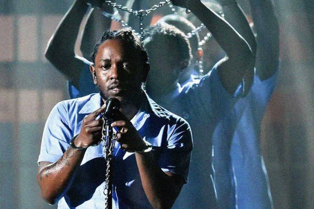 A man in a blue shirt holds a microphone with chains wrapped around his hands, while people behind him also hold up chains, dramatically lit on stage.