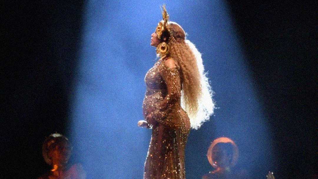 A woman in a gold, sparkling gown and ornate headpiece stands in profile under a spotlight on stage, holding a microphone, with her pregnant belly visible. Figures in gold outfits are seen blurred in the background.