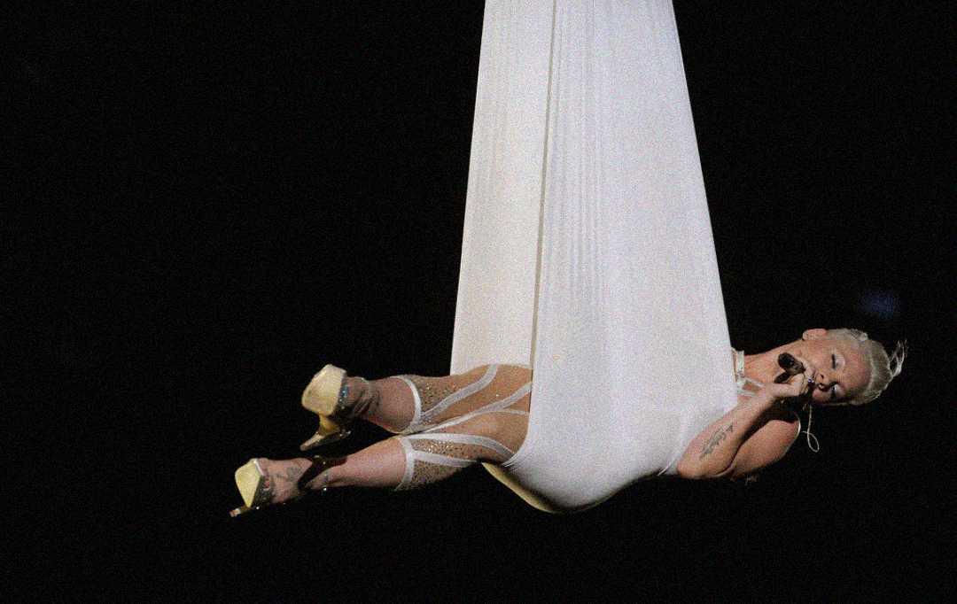 A performer in a sheer bodysuit and platform heels sings into a microphone while suspended in midair, wrapped in flowing white fabric against a dark background.