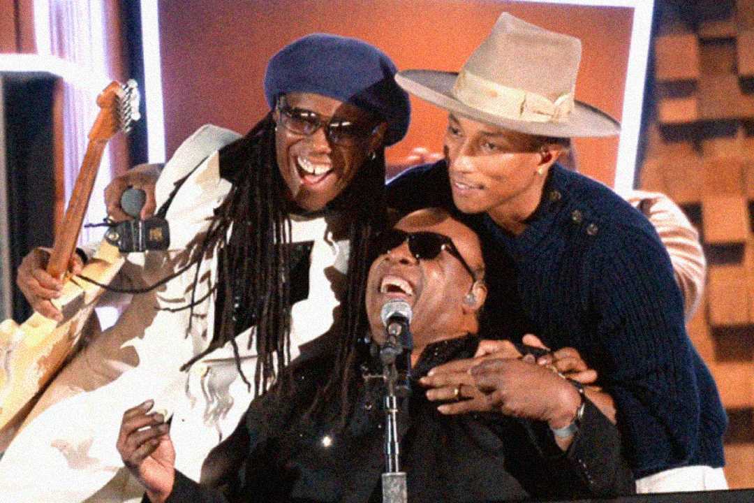 Three musicians smile and embrace on stage; one holds a guitar, another wears sunglasses and sits at a microphone, and the third wears a wide-brimmed hat, creating a joyful, celebratory moment.