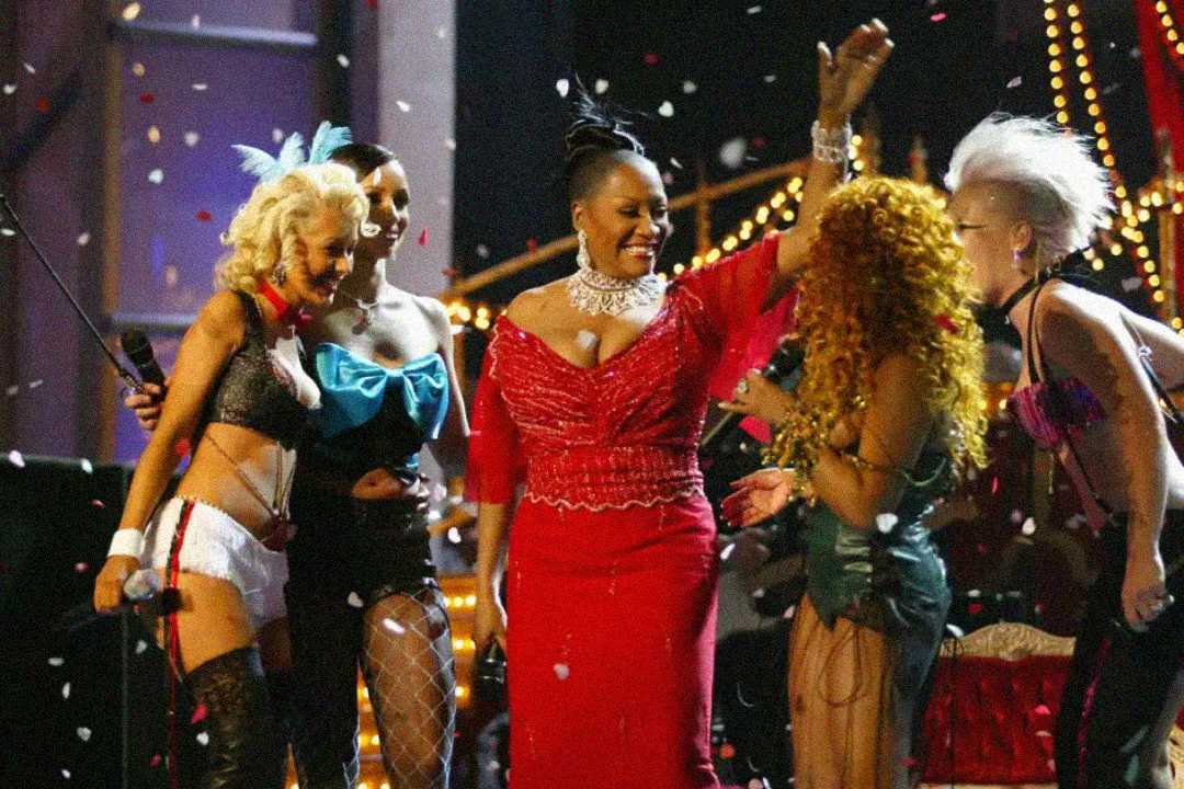 Four performers stand on stage amid falling confetti. A central figure in a red dress raises her arm, while others in colorful, elaborate costumes smile and celebrate around her under stage lights.