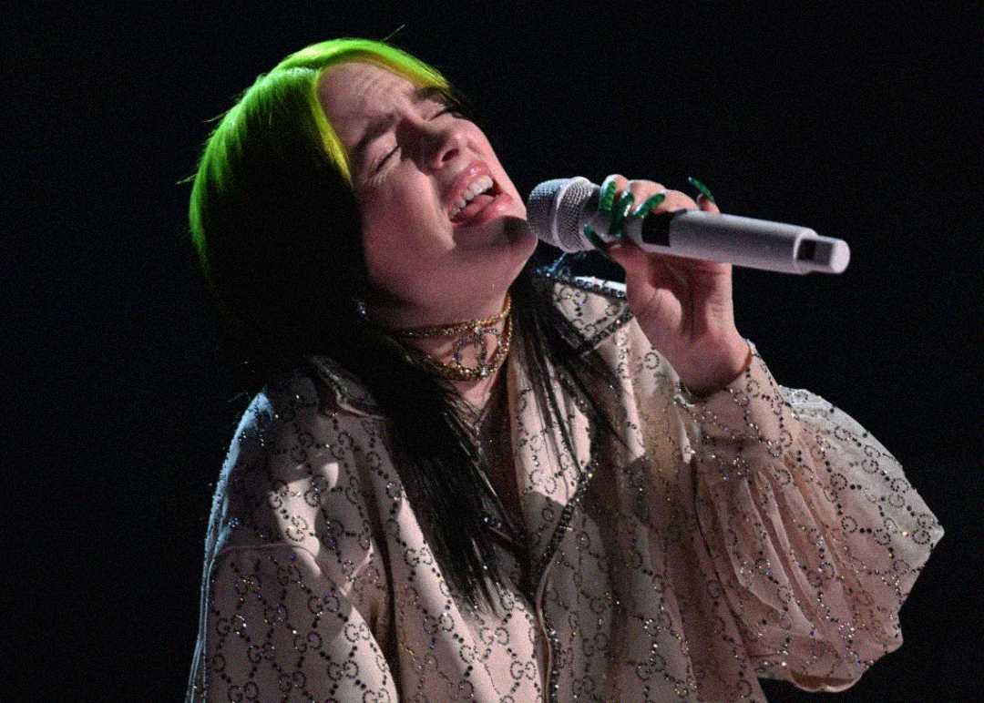 A singer with black and neon green hair passionately performs into a microphone, wearing a light-colored, patterned blouse under stage lighting.