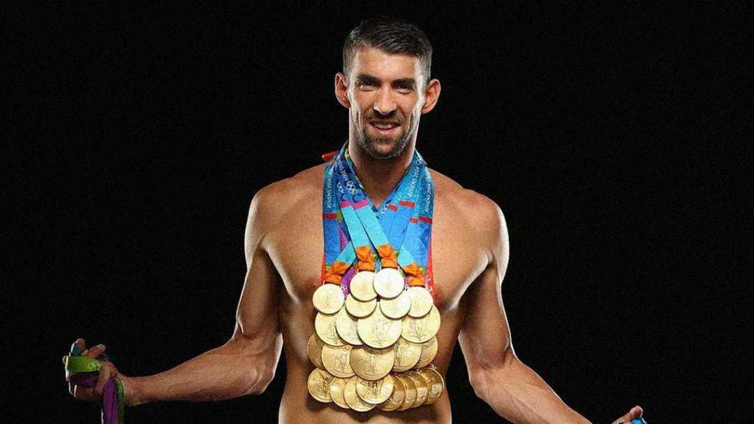 A smiling male swimmer stands shirtless against a black background, wearing numerous gold medals around his neck and holding more medals in both hands.