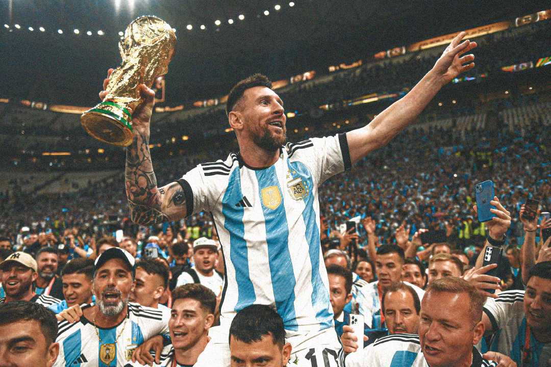 A soccer player in an Argentina jersey holds up the World Cup trophy and smiles, surrounded by jubilant teammates and fans celebrating in a packed stadium.