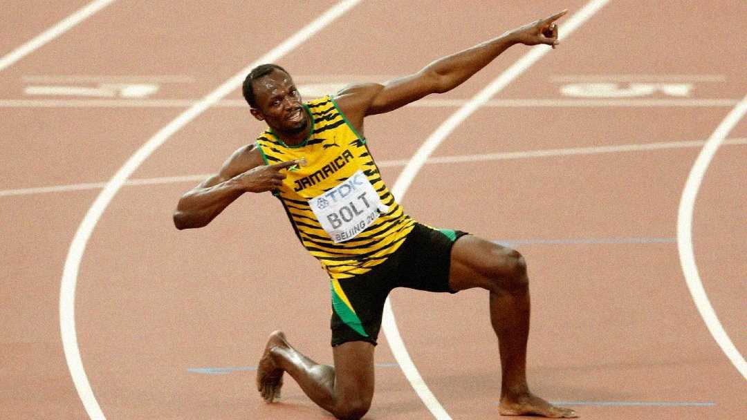 A sprinter in a yellow and green "Jamaica" uniform strikes a celebratory pose on a running track, kneeling with one arm pointed upward and the other across his chest, smiling.