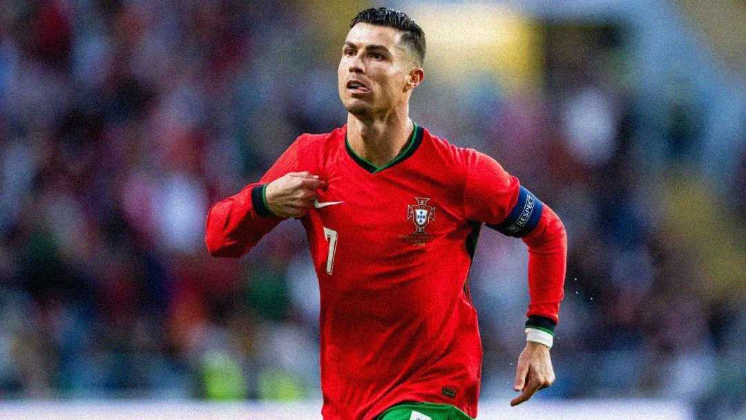 A soccer player in a red Portugal jersey with the number 7 celebrates on the field, holding his hand to his chest, with blurred spectators in the background.