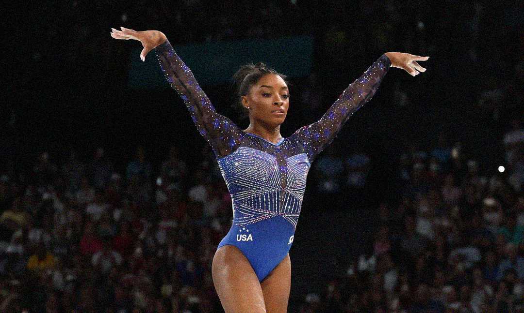 A female gymnast in a sparkling blue leotard stands confidently with arms raised during a performance. The arena behind her is filled with spectators, and the letters "USA" are visible on her outfit.