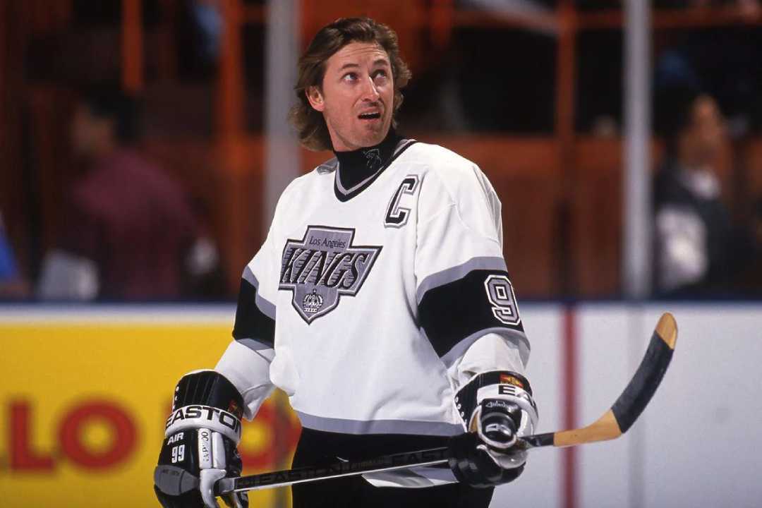 A hockey player in a white Los Angeles Kings jersey with the number 99 and a captain's "C" patch stands on the ice holding a hockey stick, looking upwards during a game.