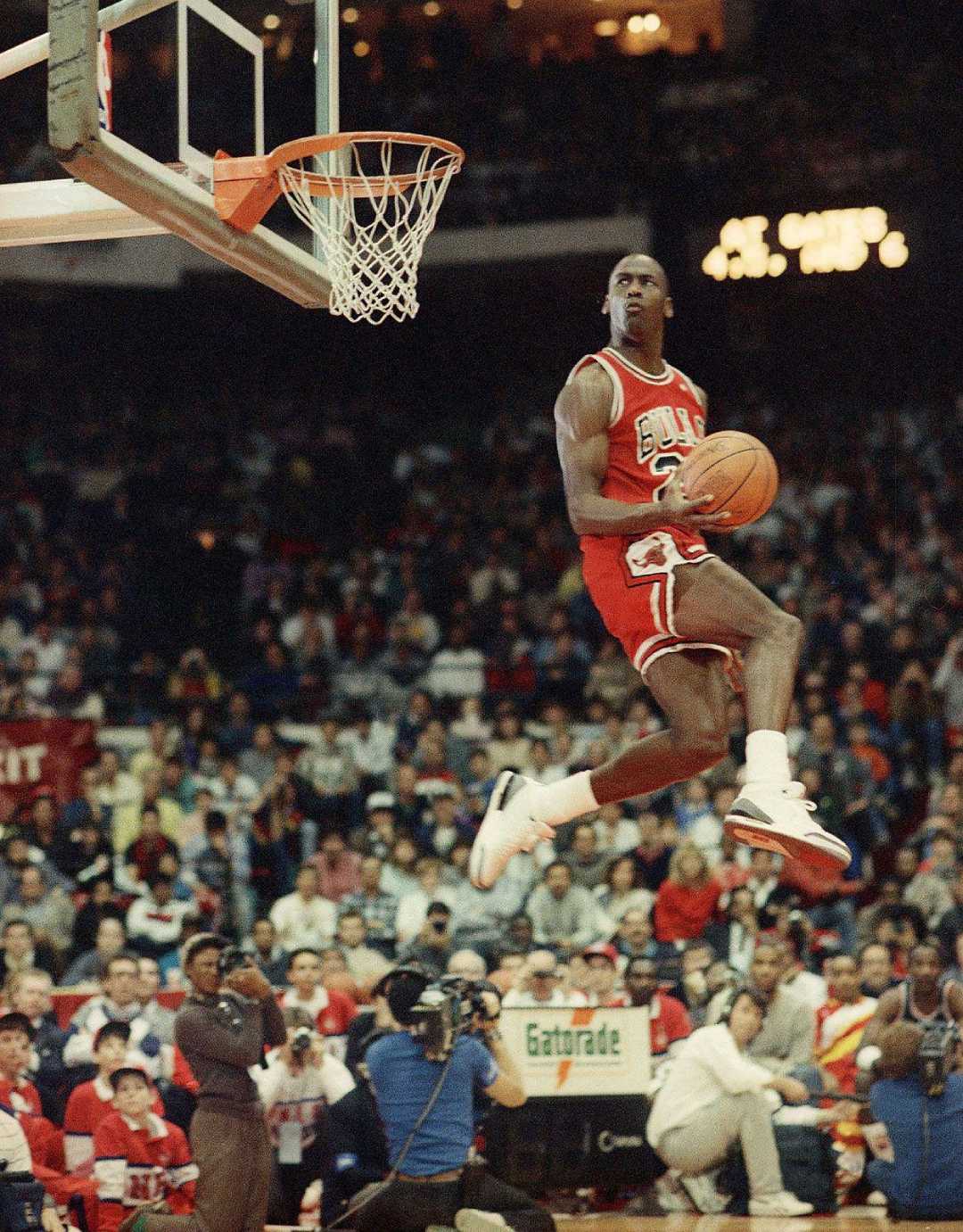 A basketball player in a red Chicago Bulls uniform is mid-air, holding a basketball under one arm as he heads toward the hoop. The crowded arena watches in anticipation.
