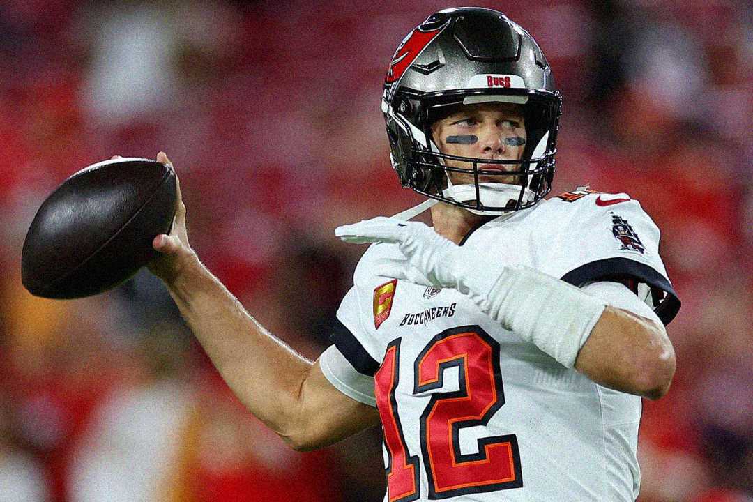 A football player wearing a Tampa Bay Buccaneers uniform and helmet, with the number 12 on his jersey, prepares to throw a football during a game. The background is blurred with red stadium seats.