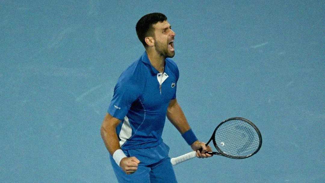 A male tennis player in a blue outfit celebrates passionately on a blue tennis court, holding his racket tightly in one hand with his mouth open in a triumphant yell.