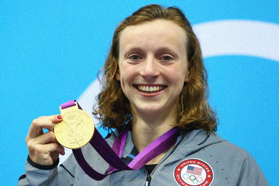 A smiling athlete wearing a gray USA Olympic jacket holds up a gold medal with a purple ribbon in front of a blue background.