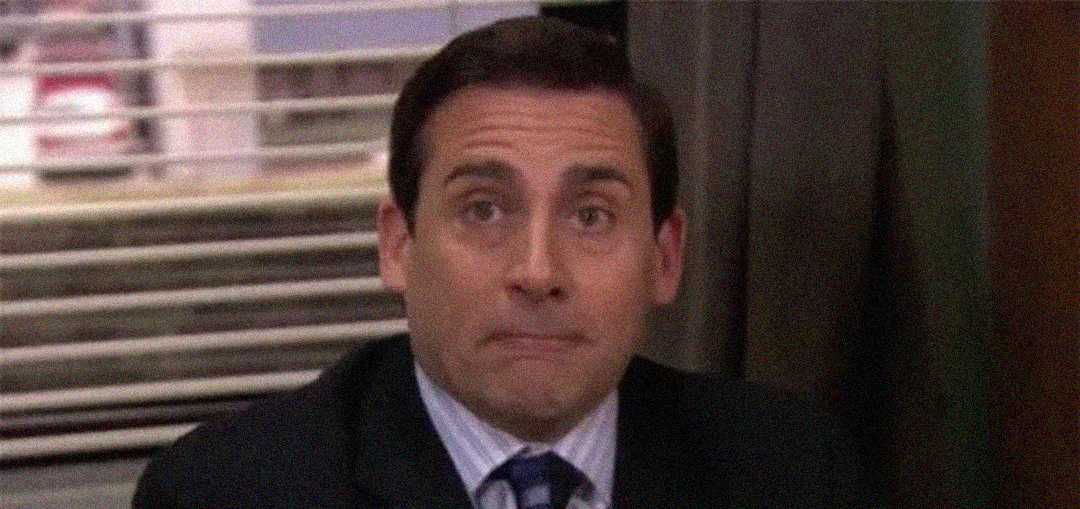 A man in a suit sits indoors, looking slightly surprised or awkward, with his lips pressed together and eyes wide open. There are blinds and office furniture visible in the background.