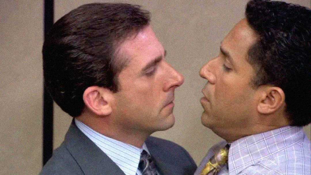 Two men in business attire stand very close, almost nose-to-nose, with serious expressions. The background suggests an office setting.