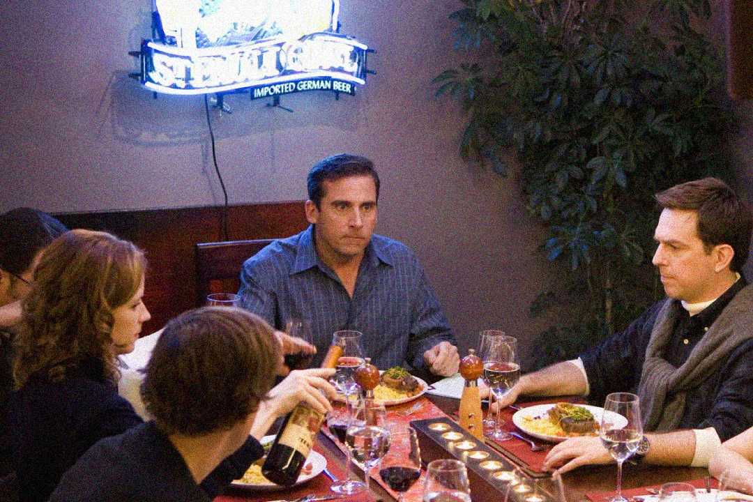 Four people sit around a dinner table with plates of food and glasses of wine. One person in the center looks serious under a bright neon beer sign on the wall. The atmosphere seems tense.
