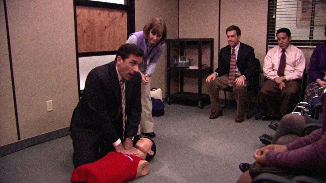 A man in a suit performs CPR on a training mannequin while others watch in a small office room. A woman supervises closely, and three seated people observe the demonstration.