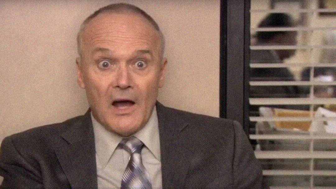A middle-aged man in a gray suit and tie sits indoors, looking shocked with wide eyes and an open mouth. Behind him is a window with blinds and an office setting visible through the glass.