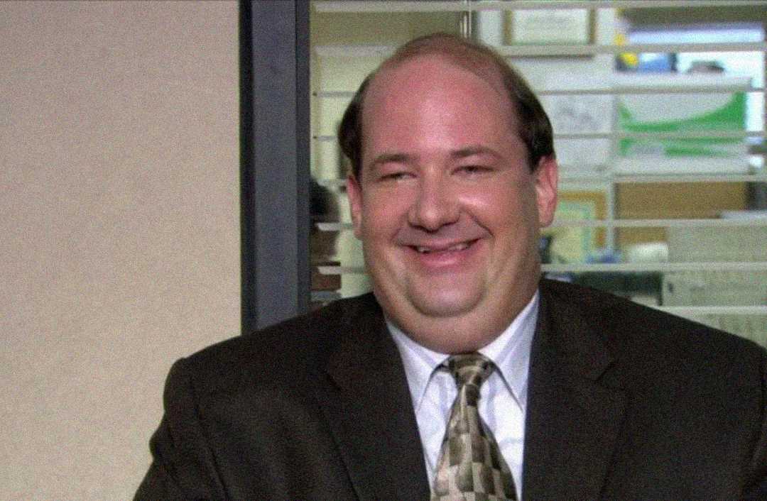 A man with thinning hair, wearing a brown suit and patterned tie, smiles while sitting in an office with glass partitions and desks in the background.
