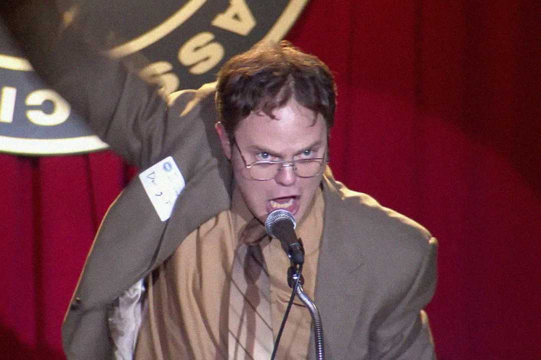 A man in a suit and glasses passionately speaks into a microphone on stage, raising one arm. He has a name tag on his jacket, and a red curtain and circular sign are visible in the background.