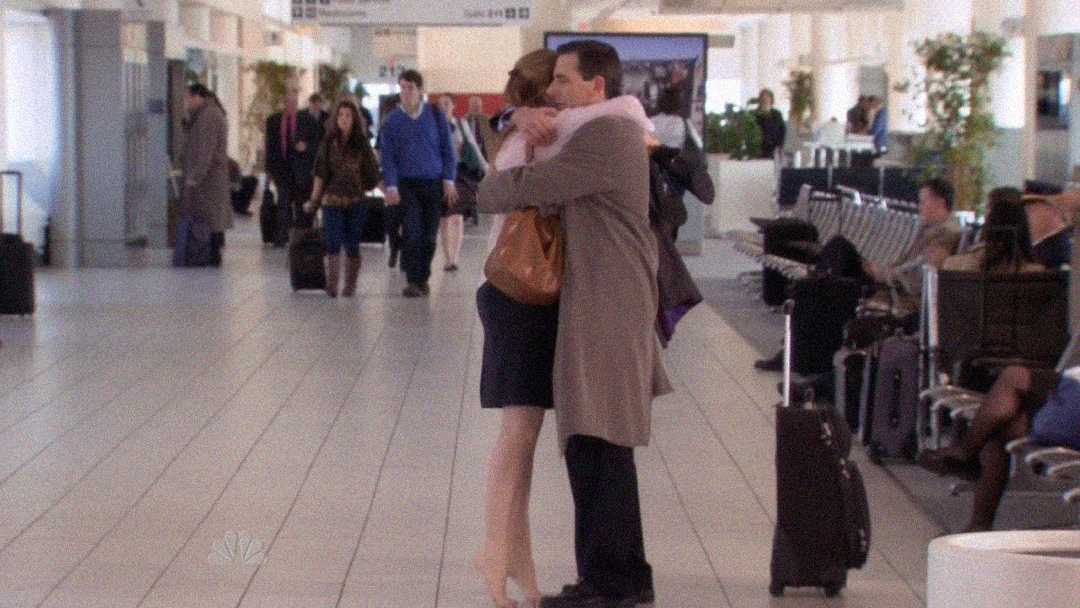 A man and woman hug tightly in an airport terminal, surrounded by people walking and sitting with luggage. The space is bright with natural light and rows of chairs line the sides.